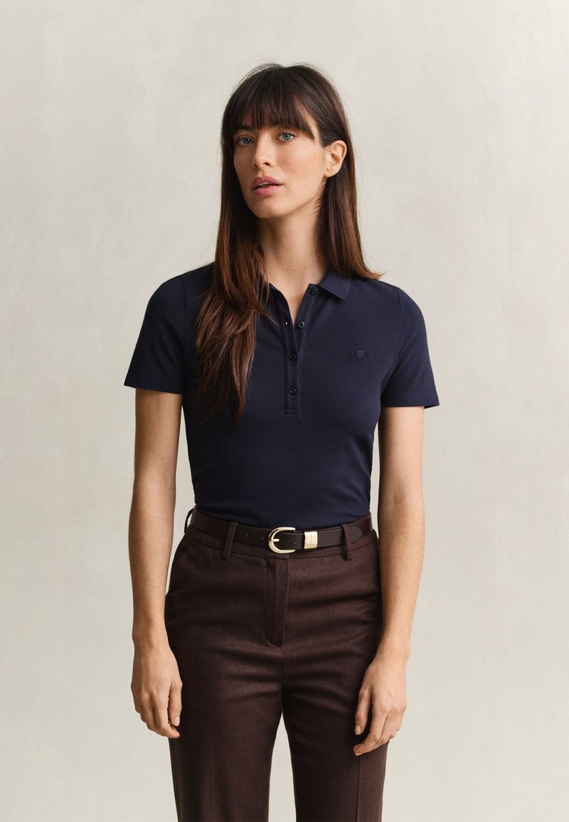 Femme aux longs cheveux bruns portant un polo bleu marine rentré dans un pantalon marron avec une ceinture noire, debout devant un fond uni.