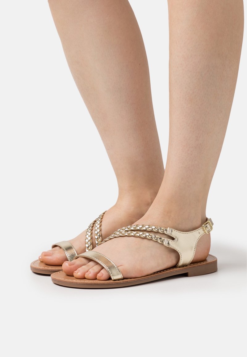 Sandales en cuir doré avec des straps tressés, bout ouvert et une boucle ajustable à la cheville. Semelle plate avec des détails marron clair.