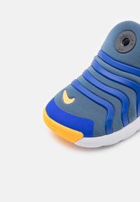 Zapatilla deportiva azul con upper texturizado gris, acentos amarillos y diseño azul ondulado. Presenta un logo circular y suela de goma.
