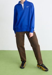 Blå, zippered sweatshirt med hög krage och vitt logotyp, kombinerat med bruna leopardmönstrade byxor och svarta sneakers med gula accenter.