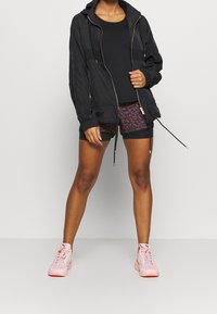 Svart lättviktsjacka med huva, kombinerad med en figurnära svart topp och mönstrade shorts. Rosa träningsskor fullbordar outfiten.