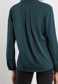 Blusa a maniche lunghe in teal con design a schiena aperta, texture morbida e dettagli con bottoni ai polsini, abbinata a legging neri.