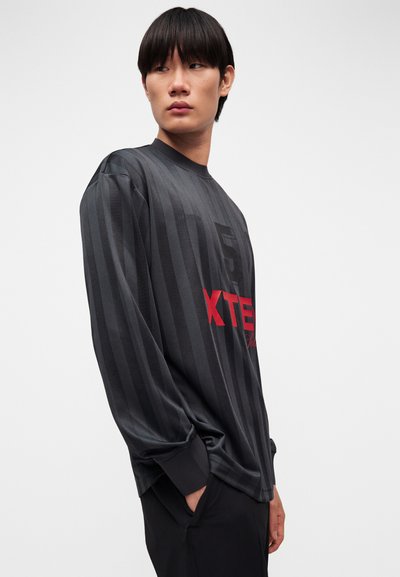 Νεαρός άνδρας που φοράει ένα χαλαρό πουκάμισο με μακριά μανίκια, μαύρο και σκούρο γκρι με κάθετες ρίγες, με κόκκινα γράμματα, στέκεται με τα χέρια στις τσέπες, κοιτάζοντας προς τα αριστερά.