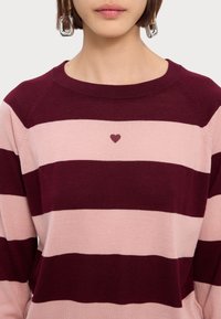 Pull rayé bordeaux et rose pâle, avec un col rond et une petite broderie en forme de cœur sur la poitrine. Texture tricotée lisse.