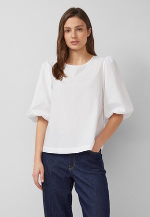 Vrouw met halflang bruin haar, gekleed in een witte blouse met pofmouwen en donkerblauwe spijkerbroek, tegen een effen grijze achtergrond.