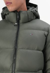 Pufferjack in olijfgroen, met een hoge kraag, zwarte rits en elastische trekkoorden. Kleine logo detail op de borst.
