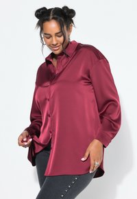 Blusa de satén burdeos con un cierre de botones en la parte delantera, mangas largas y un corte relajado. Presenta un cuello clásico y una textura suave.