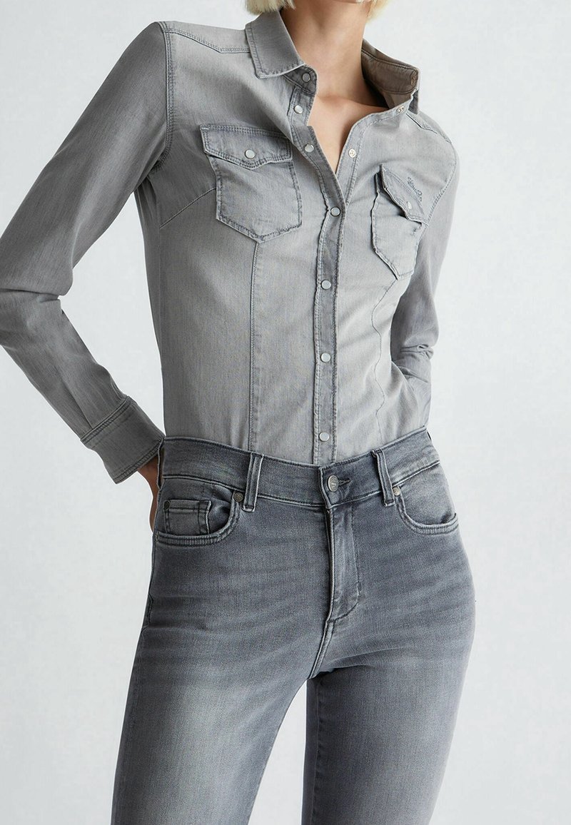 Femme portant une chemise en jean grise claire ajustée avec deux poches poitrine et un jean gris assorti, mains placées derrière les hanches.
