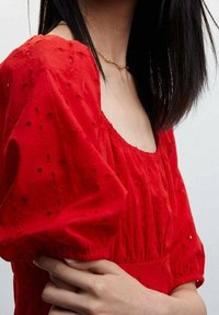 Robe en coton rouge avec des manches volumineuses, ornée de broderies en œillets et d'un décolleté carré. Texture lisse et détail de taille froncée.