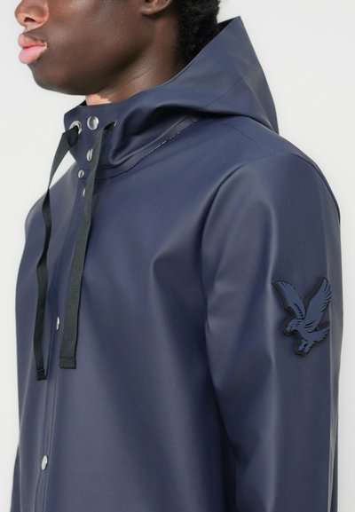 Navy wasserfeste Jacke mit hohem Kragen, Kordelzügen und Druckknopfverschlüssen. Mit einem schwarzen Adler-Patch am linken Ärmel.