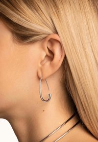 Orecchino a cerchio in argento con una forma fluida a goccia e un design slanciato. Presenta una superficie liscia e lucida che riflette la luce.