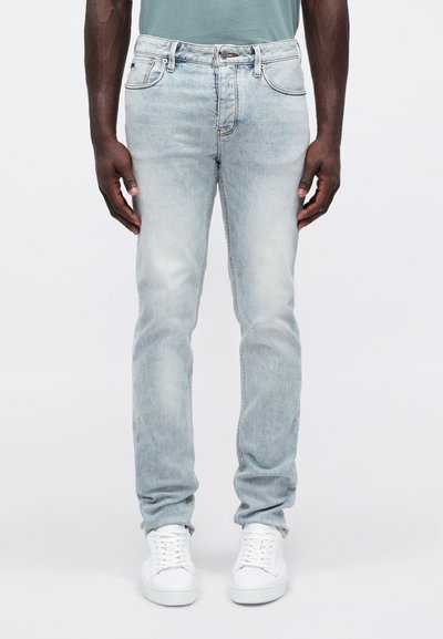 Mann iført lyseblå denimjeans, hvite sneakers, står med armene avslappet langs siden mot en enkel bakgrunn.
