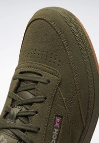 Primo piano di una sneaker in suede verde oliva con lacci, punta perforata, suola testurizzata e un piccolo dettaglio con la bandiera britannica.