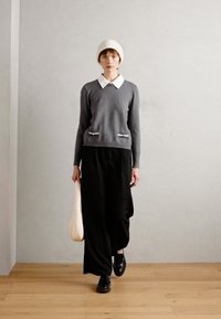 Grauer Pullover mit weißem Kragen, zwei Reißverschlusstaschen, kombiniert mit schwarzen, locker sitzenden Hosen und schwarzen Schuhen, hält eine cremefarbene Handtasche.