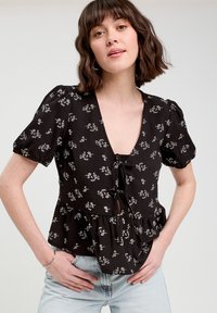 Blouse noire à motifs floraux avec des manches courtes bouffantes, décolleté en V profond, fermeture à nouer à l'avant, ourlet peplum froncé, avec des motifs floraux blancs.