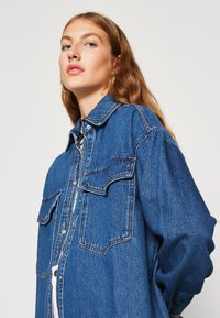 Denim srajca v srednje modri barvi, z ovratnikom, dvema sprednjima žepoma s poklopcema in sproščenim krojem, ki vključuje vidne šive.