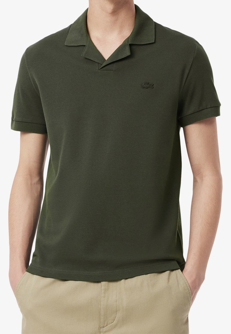Homme portant un polo à manches courtes vert foncé avec col et pantalon beige, mains dans les poches, torse visible au-dessus de la taille.