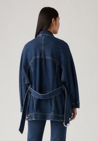 Jachetă din denim de culoare albastru închis, cu o croială lejeră, cu talie legată, mâneci supradimensionate și cusături de contrast pe șuvițe.