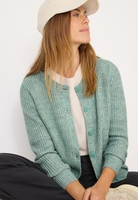 Cardigan tricoté vert clair avec un motif texturé, décolleté rond et boutons bleus clairs, associé à un haut clair et un pantalon foncé.