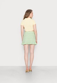 Polo amarelo de mangas curtas combinado com uma saia mini verde com estampado floral e acabamento com babados. A modelo usa sandálias amarelas de salto.