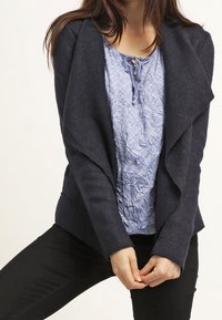 Veste bleu marine foncé avec un col drapé, en tissu doux. Portée sur un chemisier bleu clair texturé, complétée par un pantalon noir.