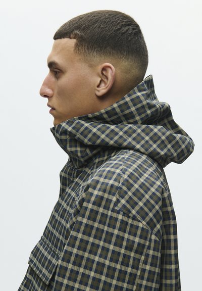 Profilo laterale di un uomo che indossa una giacca a quadri blu e beige con colletto alto, capelli corti e un'espressione neutra.