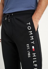 Czarne spodnie dresowe z bawełny, z białym haftowanym napisem "TOMMY HILFIGER EST. 1985" na lewej nodze, z czerwonymi i niebieskimi akcentami.