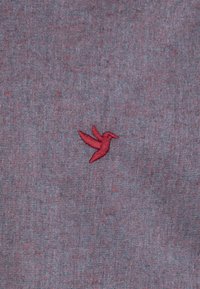 Colibrí rojo bordado sobre un fondo de tela gris texturizado con sutiles variaciones de color, creando una apariencia de tejido.