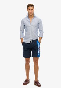 Chemise à rayures bleu et blanc ; associée à un short de bain bleu marine avec un accent bleu et du texte. Des chaussures bateau marron complètent le look.