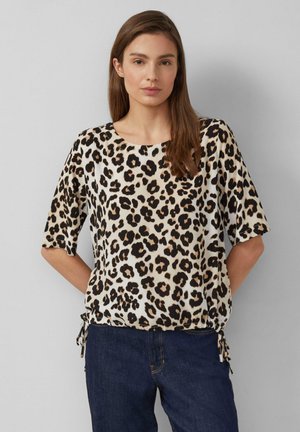 Femme portant un haut beige à motif léopard avec des manches mi-longues et un jean bleu, debout devant un fond gris clair.