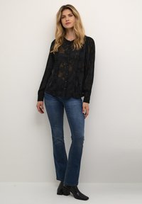 Blusa nera trasparente con motivo floreale e dettagli in balza, chiusura frontale con bottoni, abbinata a jeans blu e stivaletti neri alla caviglia.