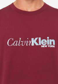 Close-up of a maroon T-shirt with white and light blue "Calvin Klein New York" logo printed across the chest.