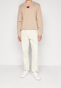 Felpa beige a girocollo con una toppa con logo rosso, abbinata a pantaloni bianchi a sigaretta e sneakers bianche. Tessuto morbido, design casual.