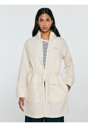 Kobieta w beżowej kurtce na zamek z troczkami i dużymi przednimi kieszeniami, pod spodem w paski koszulka, stoi na jednolitym tle.