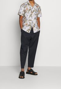 Kortärmad blommig skjorta med vit bakgrund; mörka randiga byxor; svarta lädersandaler med öppen tå och dekorativa detaljer.