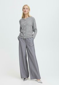 Grau gestrickte Strickjacke mit rundem Halsausschnitt, vorderem Knopfverschluss und langen Ärmeln. Kombiniert mit grauen weiten Hosen und silbernen Absätzen.