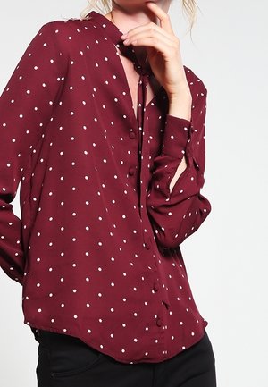 Femme portant un chemisier bordeaux à manches longues avec des pois blancs, boutonné devant et un nœud au cou, posant avec une main près du visage.