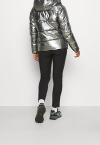 Personne portant une doudoune argentée brillante, des leggings noirs, des chaussettes blanches et des chaussures de sport noires marchant de dos sur un fond uni.