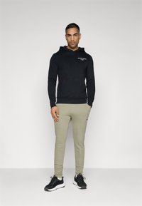 Moleton preto com capuz, bolso frontal tipo canguru e logotipo; combinado com calças de treino tapered em verde oliva claro e sapatilhas atléticas pretas.