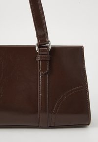 Borsa in pelle marrone scuro con manico superiore fissato da anelli in metallo argento e cuciture decorative nell'angolo in basso a destra.