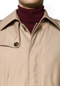 Beżowy trencz o miękkiej teksturze i z zapięciem na guziki, nałożony na bordowy prążkowany golf. Zbliżenie na kołnierz i detale materiału.