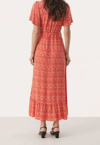 Maxikleid in leuchtend Rot mit abstraktem weißen Muster, kurzen Ärmeln, elastischem Taillenbund und gestuftem Rüschensaum. Kombiniert mit schwarzen Absätzen.
