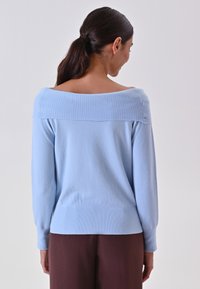Maglione azzurro chiaro con scollo a barchetta a costine, maniche lunghe e corpo testurizzato, abbinato a pantaloni marroni.