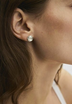 Zbliżenie na ucho kobiety z małym srebrnym kolczykiem w kształcie róży na tle brązowych włosów i jasnej skóry.