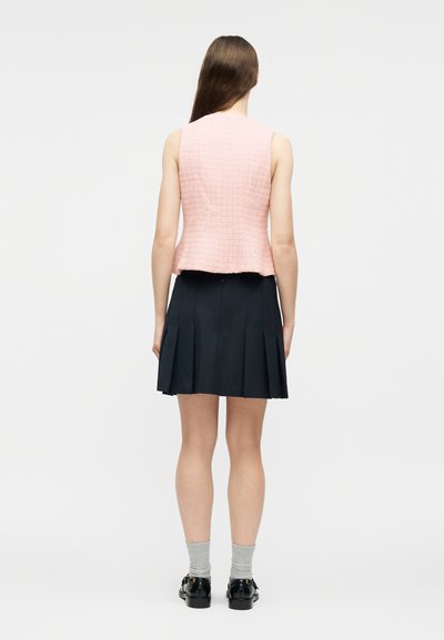 Γυναίκα με μακριά καστανά μαλλιά, γυρισμένη πλάτη, φοράει ροζ αμάνικο στρωματοποιημένο τοπ, μαύρη πλισέ φούστα, γκρι κάλτσες και μαύρα παπούτσια.