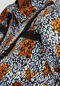 Camisa estampada con un patrón abstracto en colores naranja, negro, blanco y verde. Detalle de encaje negro notable a lo largo de las costuras.