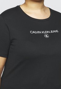 Černé bavlněné tričko s kulatým výstřihem, s bílým nápisem a logem "CALVIN KLEIN JEANS" umístěným na hrudi. Krátké rukávy.
