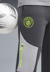 Sivé športové nohavice s svetlosivým panelom a neónovo zeleným zipsom. Na stehne sa nachádza logo Manchester City. Drží čierno-biely futbal.