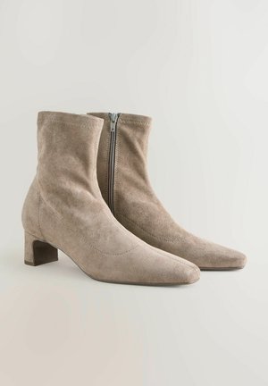Beige ruskindsankelstøvler med spidse tæer, kort blokhæl og lynlåsdetalje på siden. Blød tekstur og enkel, glat design.