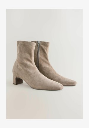 Botine din suede bej, cu vârf ascuțit, toc scurt și detaliu cu fermoar pe lateral. Textură moale și design simplu, neted.